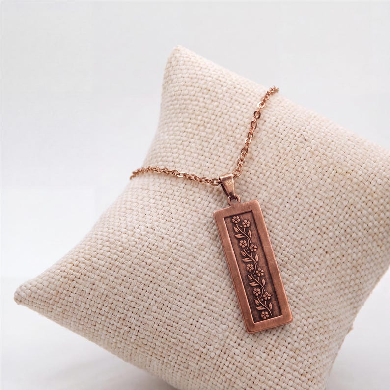 Puede incluir: Un collar con colgante rectangular de tono cobre que presenta un dise&ntilde;o de enredadera floral en relieve. El colgante cuelga de una cadena de metal a juego, mostrando una disposici&oacute;n vertical de peque&ntilde;as flores y hojas dentro de un borde hundido.