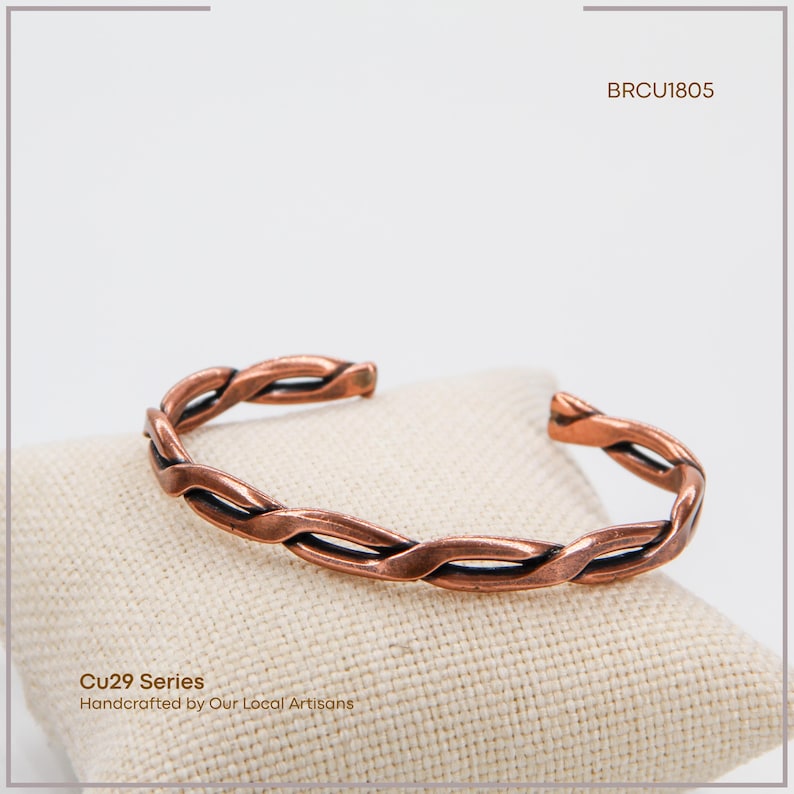 K&ouml;nnte beinhalten: Ein kupferfarbenes Armband mit geflochtenem Design. Das Armband wird auf einem beigen Stoff pr&auml;sentiert. Der Text "BRCU1805" befindet sich in der oberen rechten Ecke. Der Text "Cu29 Series Handcrafted by Our Local Artisans" befindet sich in der unteren linken Ecke.