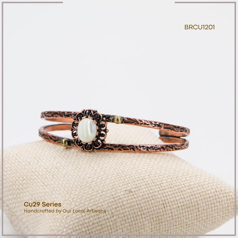 Puede incluir: Brazalete de pu&ntilde;o cobrizo con una piedra ovalada blanquecina en el centro. El brazalete presenta un dise&ntilde;o floral alrededor de la piedra y una textura martillada. Peque&ntilde;os detalles dorados se colocan a lo largo de la banda. El texto "Cu29 Series" y "Handcrafted by Our Local Artisans" es visible.