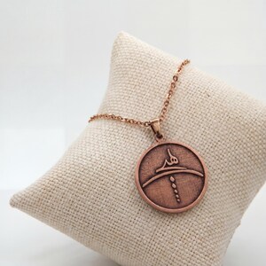 Op de afbeelding: Een koperkleurige hangerketting met een cirkelvormige bedel met een gegraveerd ontwerp. De bedel hangt aan een delicate ketting en rust op een beige kussen. De hanger heeft een Arabische inscriptie en een gestileerd ontwerp.