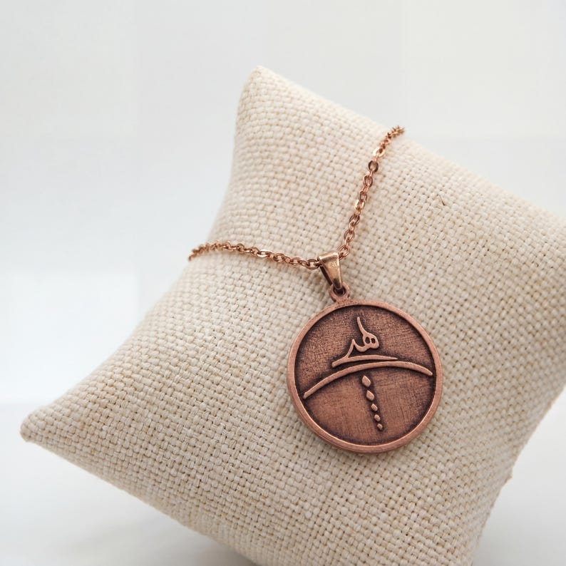 Op de afbeelding: Een koperkleurige hangerketting met een cirkelvormige bedel met een gegraveerd ontwerp. De bedel hangt aan een delicate ketting en rust op een beige kussen. De hanger heeft een Arabische inscriptie en een gestileerd ontwerp.
