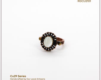 Può includere: Un anello color rame con una pietra ovale bianco sporco al centro, circondata da un design a filigrana scura. L'anello presenta piccoli accenti decorativi sulla fascia. Il testo "Cu29 Series" e "Handcrafted by Our Local Artisans" è visibile.