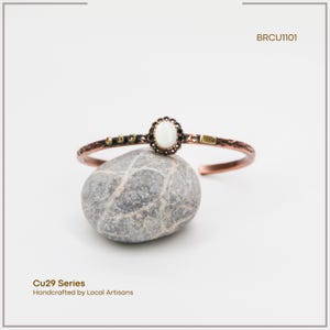 Pu&ograve; includere: Bracciale a polsino in rame con una pietra ovale bianco sporco al centro e dettagli decorativi. Il bracciale fa parte della serie Cu29, realizzato a mano da artigiani locali. Il testo "BRCU1101" &egrave; visibile.