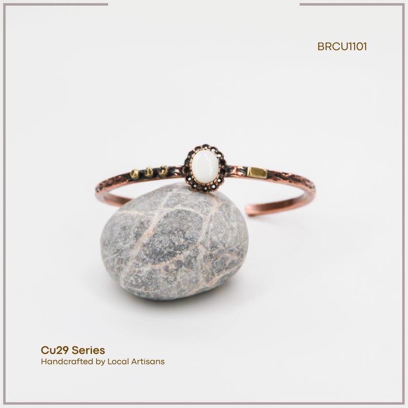 Pu&ograve; includere: Bracciale a polsino in rame con una pietra ovale bianco sporco al centro e dettagli decorativi. Il bracciale fa parte della serie Cu29, realizzato a mano da artigiani locali. Il testo "BRCU1101" &egrave; visibile.