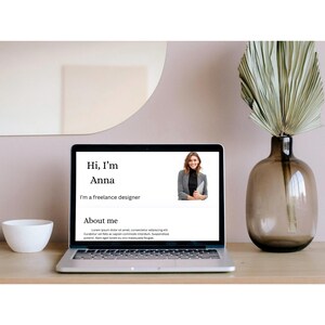 May include: A laptop displaying a website with the text "Hi, I'm Anna, I'm a freelance designer." A photo of a woman is on the screen. A white bowl and a decorative vase with dried foliage are on the wooden desk.