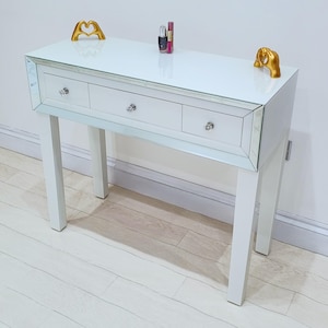 May include: White mirrored console table with three drawers and gold decorative accents. The table has a glass top and white legs.