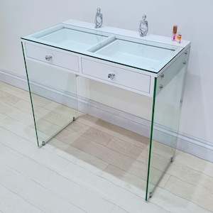 May include: White vanity table with two drawers and a glass top. The table has two small compartments on the top, and the glass top is supported by clear glass legs.