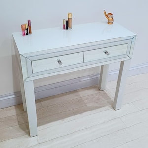 May include: A white dressing table with two drawers and mirrored detailing. The table has a high-gloss finish and crystal-style drawer knobs. Various makeup products, including lipsticks and foundation, are arranged on the surface. A small gold ornament is on the right.