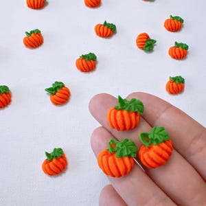 May include: Miniature orange pumpkins with green stems are scattered across a white surface. The pumpkins are round with vertical ridges and a green leaf design. Some pumpkins are held in a hand. These decorative pumpkins are perfect for fall-themed crafts.