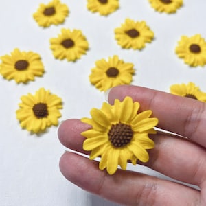 May include: A collection of yellow felt sunflower embellishments with brown centres. The sunflowers are arranged in a pattern on a white surface.