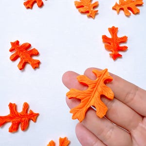 May include: Vibrant orange leaf-shaped decorations scattered on a white background. The leaves feature detailed veining and a 3D effect. One leaf is held by a hand. Suitable for crafting and seasonal decorations.