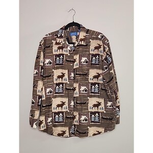 Op de afbeelding: Button-down shirt met lange mouwen met een herhalend patroon van elanden, hutten en bomen in bruine, beige en blauwe vierkanten. Het shirt hangt aan een zwarte hanger tegen een witte achtergrond.