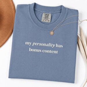 May include: A blue short-sleeved t-shirt with a crew neckline featuring the white text "my personality has bonus content" printed on the front. The shirt has a Comfort Colors brand label inside the collar and is paired with a thin silver necklace.