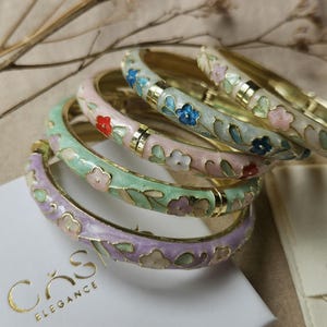 May include: A set of four hinged metal bangles featuring enamel floral patterns in pastel shades of lavender, mint green, soft pink, and cream. Each bracelet has gold-tone accents and small painted flowers. The bangles rest on a white box labeled CASI ELEGANCE.