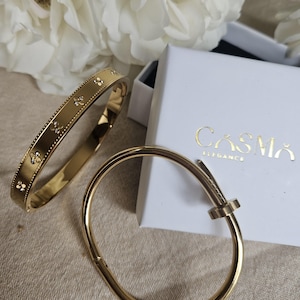 Peut inclure: Deux bracelets en métal doré et une boîte blanche avec le texte doré CASMA ELEGANCE. L'un des bracelets est un anneau plat avec un bord perlé et des motifs floraux, tandis que l'autre est un anneau lisse et arrondi avec un design en forme de clou.