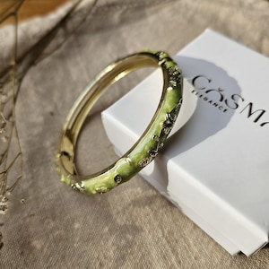 Peut inclure: Un bracelet jonc doré avec une finition en émail vert clair et des détails floraux en relief. Le bracelet est placé à côté d'une boîte blanche sur laquelle est imprimé le texte "COSMA ELEGANCE" en noir sur la surface supérieure de l'emballage.