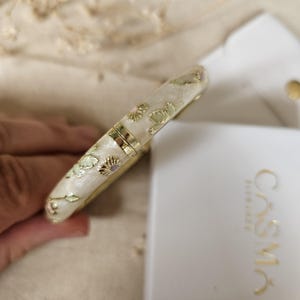 May include: A hinged bangle bracelet with a cream-colored pearlescent finish, featuring gold-tone floral and butterfly motifs. The bracelet is positioned next to a white product tag with the gold-printed text CASMA ELEGANCE.