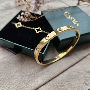 May include: A gold-coloured bangle bracelet with clear gemstones and clover-shaped cutouts. A matching gold bracelet with clover charms is displayed in a green jewellery box. The box is open, revealing a black velvet interior. The background is a neutral beige.