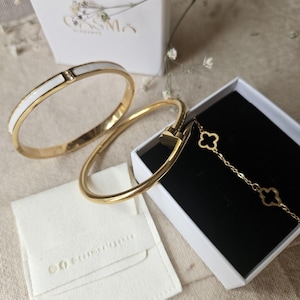 Könnte beinhalten: Zwei goldfarbene Metallarmreifen und ein goldfarbenes Kettenarmband mit kleeblattförmigen Anhängern. Ein Armreif hat eine weiße Einlage mit einem kleinen Stein. Der Schmuck wird mit einer weißen, mit Cosma Elegance beschrifteten Schachtel gezeigt.