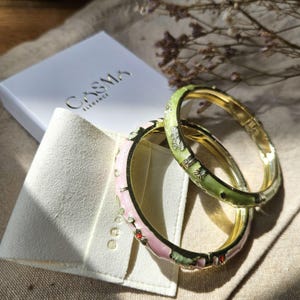 May include: Two hinged bangle bracelets with a gold-tone metal base. One bracelet features a light pink enamel finish with small floral accents, while the other has a light green enamel finish with similar floral details. A white box with the text CASMA ELEGANCE is visible.