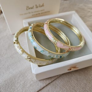 May include: Three gold bangles with floral patterns in white, pale blue, and pink enamel. The bangles are presented in a white box, with a gift box in the background that reads "Best Wish".