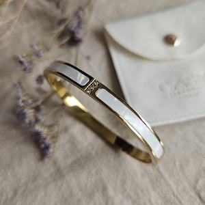 May include: A gold-toned bangle bracelet with white mother-of-pearl inlays and a small section with three small clear crystals. The bracelet has a hinged design and a clasp. A white pouch is in the background.