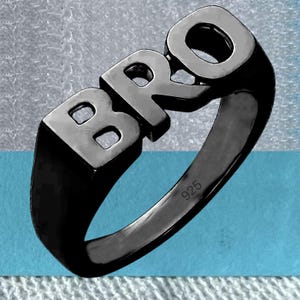 May include: A black ring featuring the word "BRO" in capital letters. The ring is made of a dark metal with a polished finish. The number "925" is engraved on the inside of the ring.