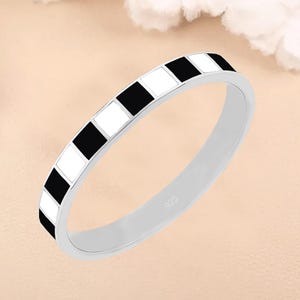 May include: A silver ring with a checkerboard pattern of black and white squares. The ring is a simple band design with a polished finish. The number "925" is stamped on the inside of the ring.