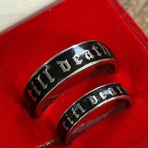 May include: Two silver rings with black enamel, each inscribed with the words "till death" in a gothic font. The rings are displayed in a red ring box, suggesting a jewelry or wedding theme.