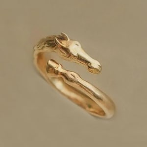 Könnte beinhalten: Ein goldfarbener Ring mit einem Pferdekopf-Design. Der Ring umschließt den Finger, wobei sich der Pferdekopf oben befindet und der Körper das Band bildet. Der Ring besteht aus einem glänzenden, metallischen Material.