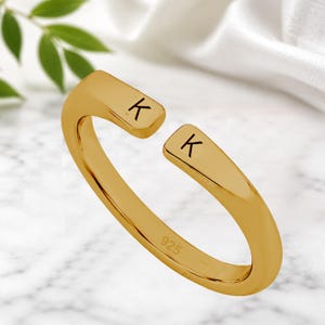 Op de afbeelding: Een goudkleurige open ring met een gepolijste afwerking en een opening aan de bovenkant. Elk uiteinde van de ring is gegraveerd met de letter K. De binnenzijde van de ring is duidelijk gestempeld met het keurmerk 925, wat duidt op sterling zilver.