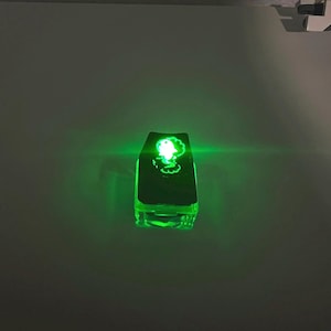 May include: A glowing green rectangular object with a stylized image of a face. The object is illuminated with green light, creating a bright, eye-catching effect. The object appears to be made of a transparent material.