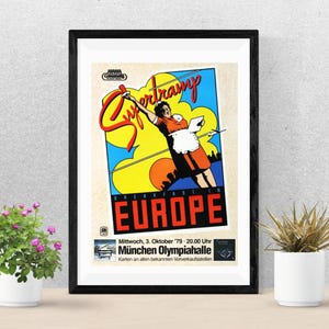 Puede incluir: Póster vintage enmarcado para la gira "Breakfast in Europe" de Supertramp. El póster presenta a una mujer con un vestido naranja y un delantal blanco, con el nombre de la banda en letras grandes y estilizadas. El póster incluye la fecha y el lugar de un concierto en Múnich.