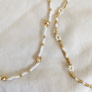 May include: Two beaded necklaces with white beads and gold flower accents. The necklaces are on a white fabric background.