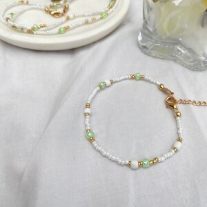 May include: A delicate white and green beaded bracelet with a gold clasp. The bracelet is made with small, round beads and has a simple, elegant design.