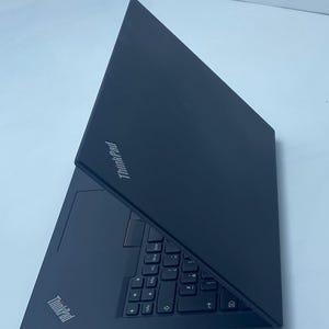 Puede incluir: Un portátil ThinkPad negro mostrado desde una perspectiva angular con la tapa parcialmente abierta. El dispositivo presenta un acabado negro mate, un teclado negro con un puntero rojo y el logotipo de ThinkPad en la tapa y el reposamuñecas.