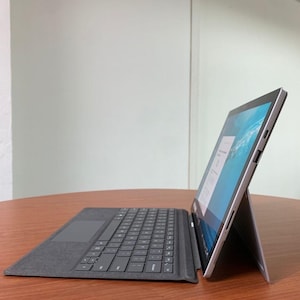 May include: A silver laptop with a black keyboard and a screen displaying a document. The laptop is propped up on a stand, and the keyboard is attached. The laptop is on a brown wooden table.