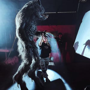May include: A large, dark gray werewolf costume with a menacing expression. The werewolf is standing on a dark floor with a person standing in front of it. The person is wearing a black shirt and ripped jeans.