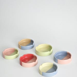 May include: Six small, round ceramic dishes, each featuring a two-tone color design. The colors include combinations of pastel pink, light blue, soft yellow, and bright red. Each dish has a smooth, glossy finish and a small notch on the rim.