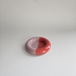 May include: A small, circular ceramic dish with a smooth, glossy finish. The object is divided into two distinct halves, with one side colored in a soft, pale lavender and the other side in a vibrant, saturated coral red.