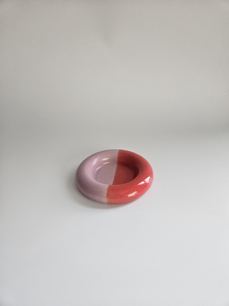 May include: A small, circular ceramic dish with a smooth, glossy finish. The object is divided into two distinct halves, with one side colored in a soft, pale lavender and the other side in a vibrant, saturated coral red.