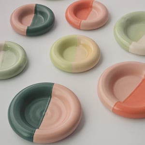 May include: A collection of small, round ceramic dishes. Each dish is divided into two color sections, with hues including green, pink, peach, and yellow. The dishes are arranged on a white surface.