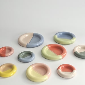 May include: A collection of small, round ceramic dishes in various sizes, each featuring a two-tone color design. The dishes display combinations of pastel and vibrant hues including pink, blue, green, yellow, and coral, with a smooth finish.