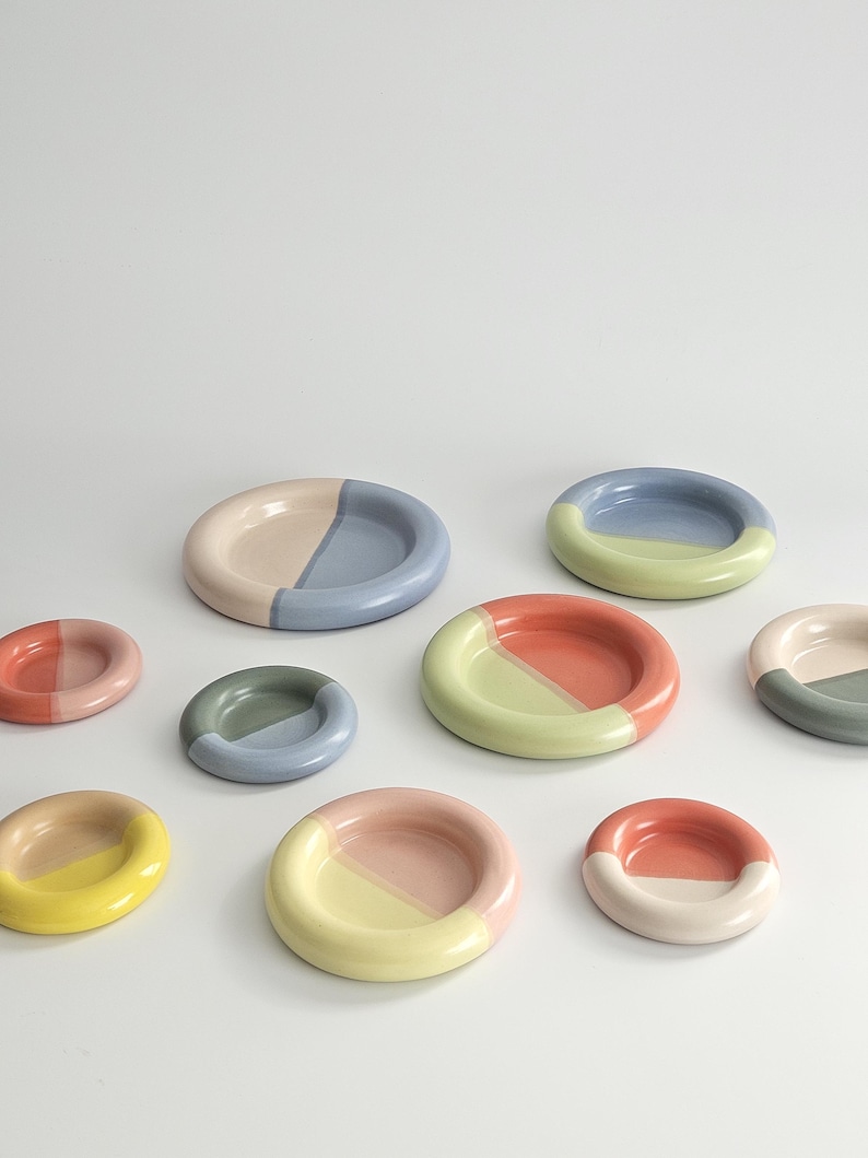 May include: A collection of small, round ceramic dishes in various sizes, each featuring a two-tone color design. The dishes display combinations of pastel and vibrant hues including pink, blue, green, yellow, and coral, with a smooth finish.