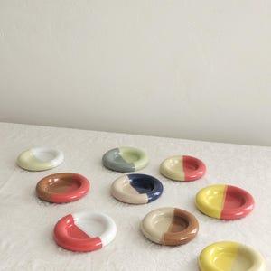 May include: A collection of nine small, round ceramic dishes arranged in a group. Each dish features a two-toned color scheme with glossy finishes, including combinations of yellow, red, white, blue, brown, and sage green in various patterns.