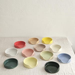 May include: A collection of twelve circular ceramic spoon rests in various solid colors including red, yellow, blue, green, pink, beige, and white. Each spoon rest features a smooth finish and a distinct cutout notch on one side of the rim.