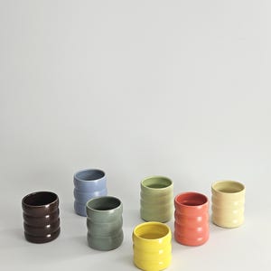 May include: A collection of seven small ceramic cups in various colors including dark brown, light blue, sage green, olive green, bright yellow, coral pink, and cream. Each cup features a distinct ribbed, cylindrical shape.