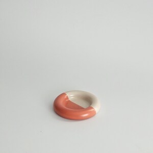 May include: A small, circular ceramic dish with a smooth, rounded rim. The object features a two-tone color scheme, with one half finished in a solid terracotta orange glaze and the other half in a neutral, off-white cream color.