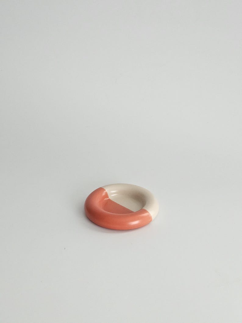 May include: A small, circular ceramic dish with a smooth, rounded rim. The object features a two-tone color scheme, with one half finished in a solid terracotta orange glaze and the other half in a neutral, off-white cream color.