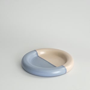 May include: A round ceramic dish featuring a smooth, glossy finish. The surface is divided into two distinct sections, with one half colored in a soft, muted blue and the other half in a creamy, off-white tone, creating a simple, balanced design.